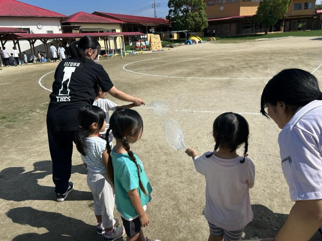 うちわでシャボン玉を作る園児をサポートする高校生